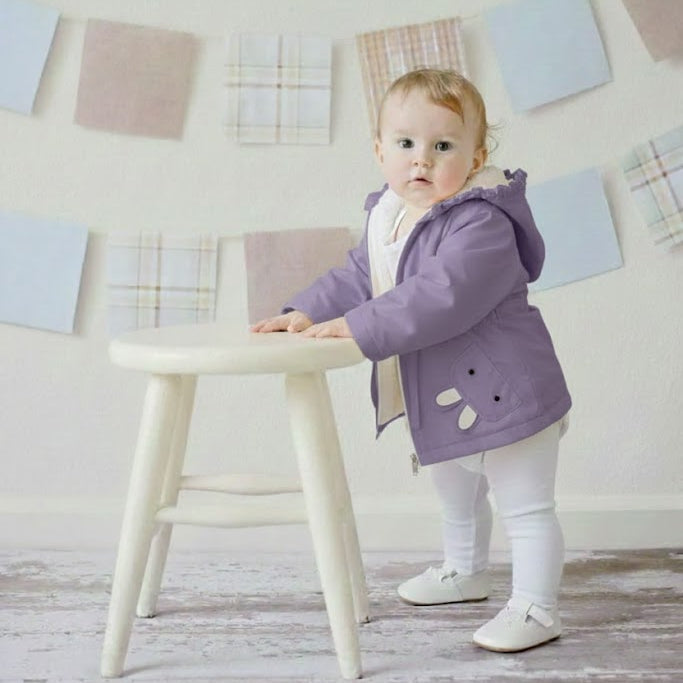 Child in a purple coat standing on a stool against a light-colored wall with decorative elements.