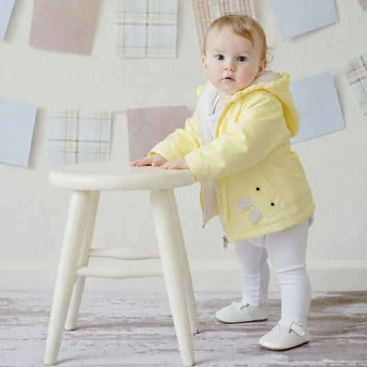 Baby in a yellow coat standing next to a white stool against a light-colored wall with decorative elements.